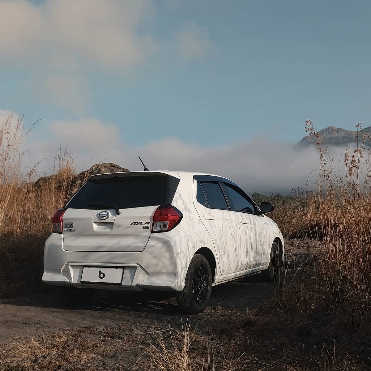 A white Toyta Agya car rests on a rustic path amidst golden grasses, with the serene Indonesian landscape of mountains and clouds visible in the distance.