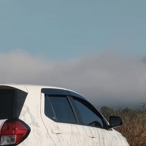 A white Toyta Agya car rests on a rustic path amidst golden grasses, with the serene Indonesian landscape of mountains and clouds visible in the distance.