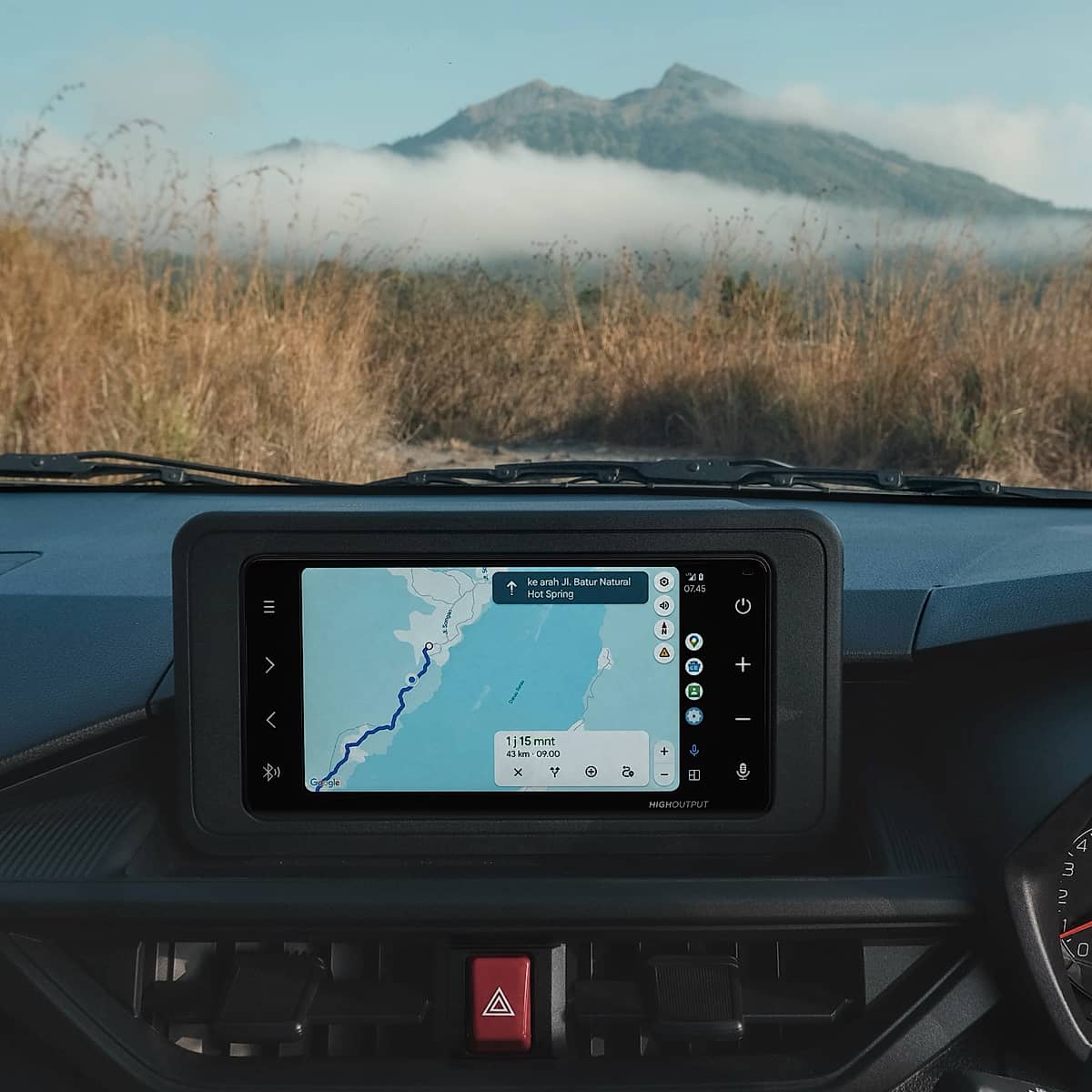 Dashboard view from inside a car, with a GPS navigation screen displaying a map of Bali and a mountain landscape visible through the windshield.