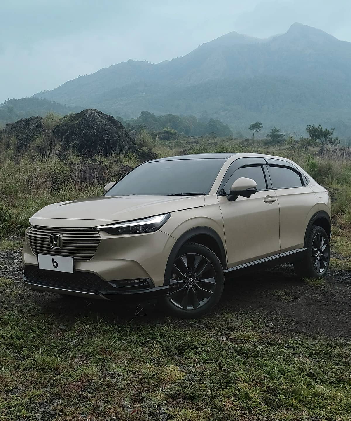 A light beige Honda HR-V SUV parked on a dirt track with green grass, rocky terrain, and a misty mountain range in the background.