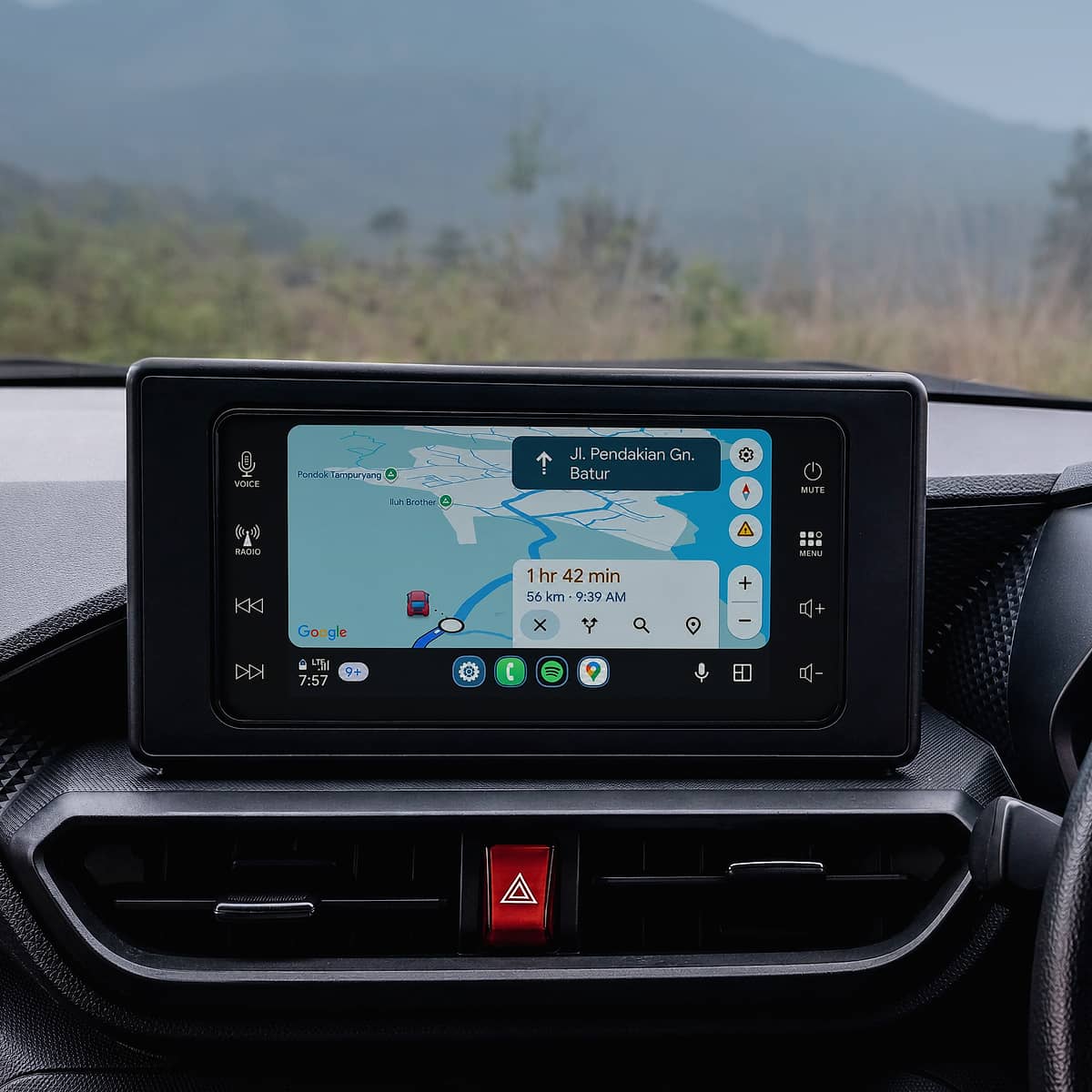 Close-up of a car's dashboard showing a navigation screen with a map and directions, with a blurred background of mountains and foliage, suggesting a scenic drive in Bali.