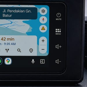 Close-up of a car's dashboard showing a navigation screen with a map and directions, with a blurred background of mountains and foliage, suggesting a scenic drive in Bali.