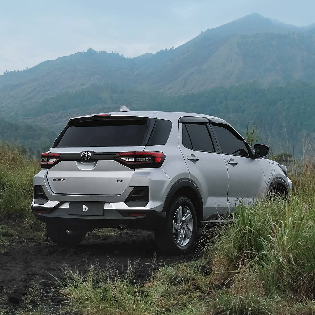Toyota Raize positioned on a rough, earthy track amidst vibrant green foliage, with majestic mountains dominating the horizon in Bali.