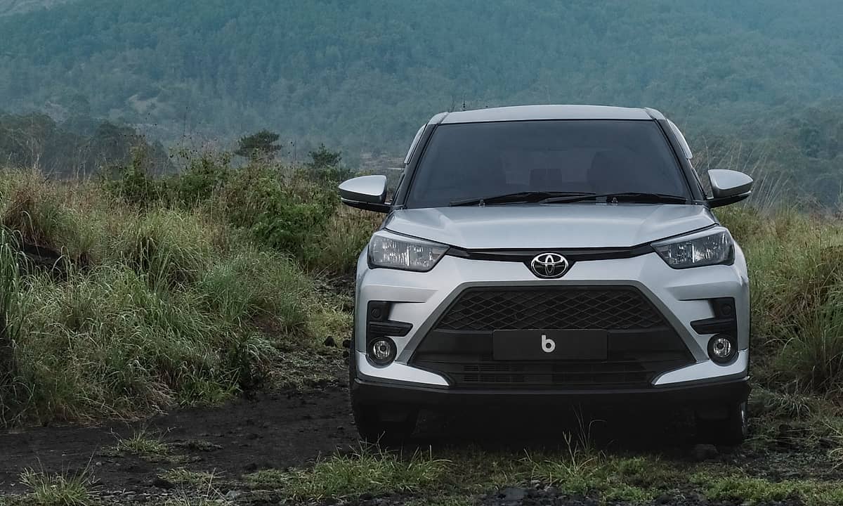 A silver Toyota Raize parked on a dirt road with lush green grass and a majestic mountain range in the background under a cloudy sky.