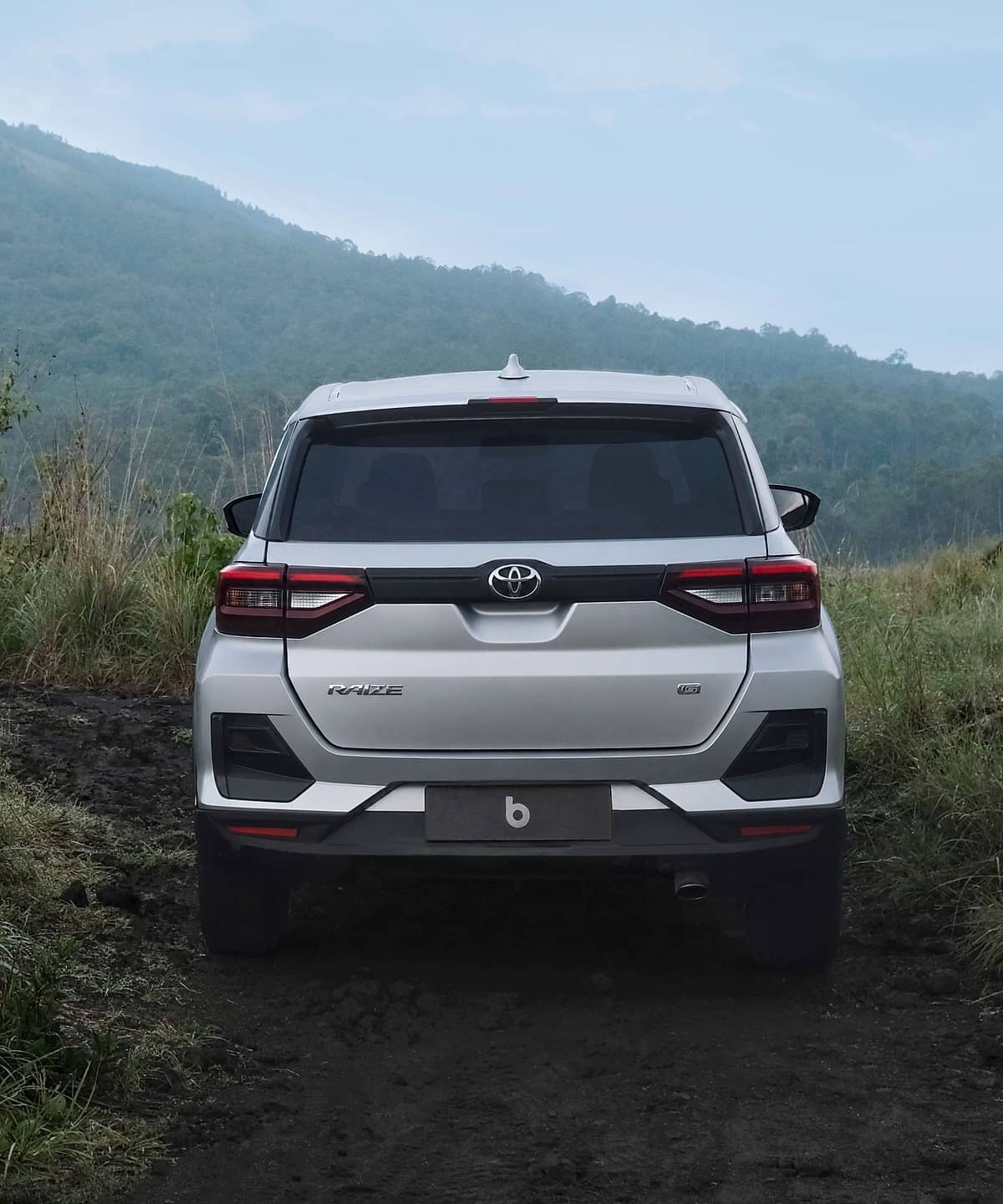 A white SUV is parked on a dusty, off-road path, surrounded by tall green grass and with a hazy mountain peak dominating the horizon.