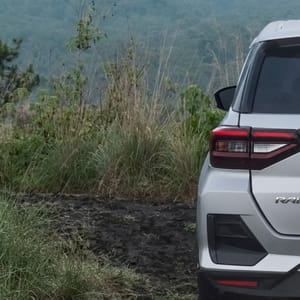 A white SUV is parked on a dusty, off-road path, surrounded by tall green grass and with a hazy mountain peak dominating the horizon.