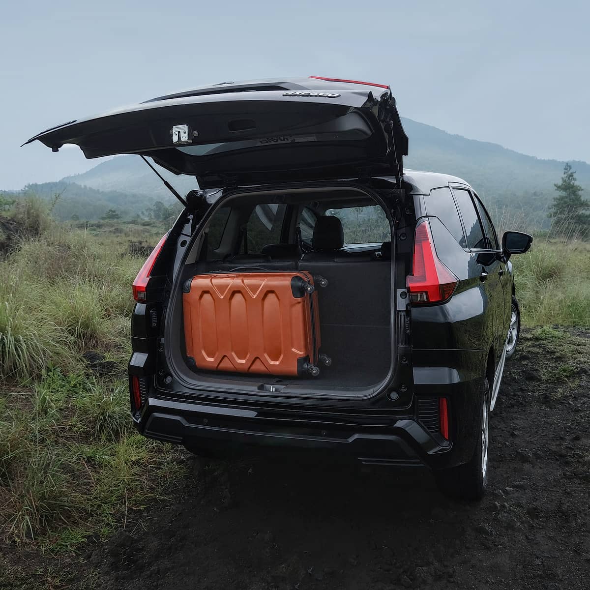 Black Mitsubishi Xpander with open trunk and orange suitcase in a rural Bali landscape.