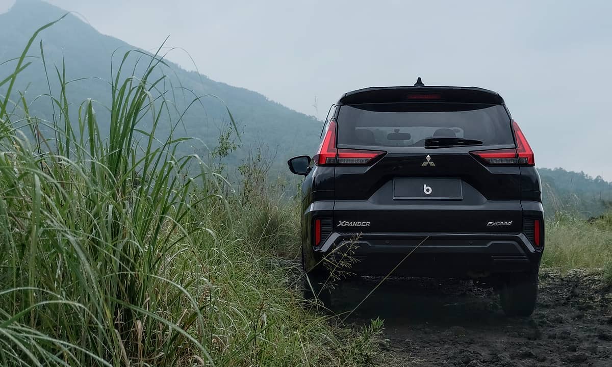 The back of a dark-colored Mitsubishi Xpander, viewed from behind, with the natural landscape of Bali featuring dense foliage and a prominent, cloud-shrouded mountain.
