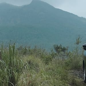 A black Mitsubishi Xpander parked on a dirt road with lush green grass and mountains in the background, cloudy sky.