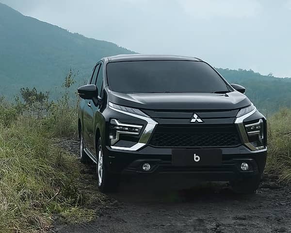 A black Mitsubishi Xpander parked on a dirt road with lush green grass and mountains in the background, cloudy sky.