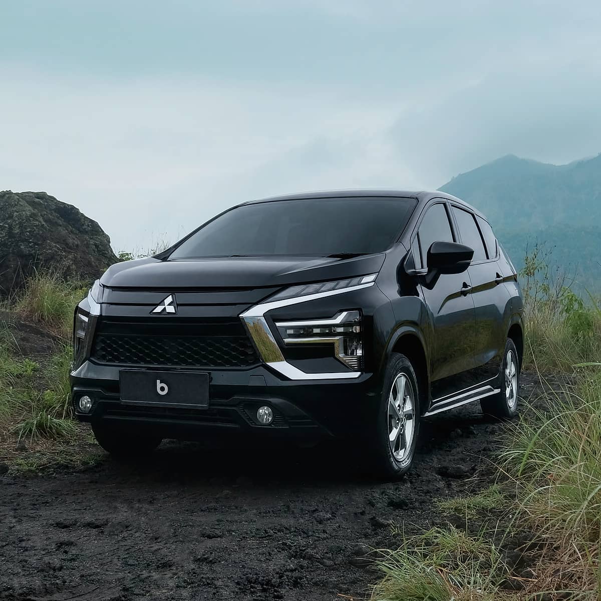 Black Mitsubishi Xpander parked on a dirt road with tall green grass and mountains in the background.