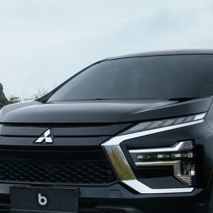 Black Mitsubishi Xpander parked on a dirt road with tall green grass and mountains in the background.