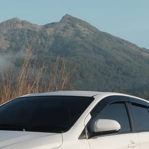 White Toyota Agya parked on dirt road with dry grass and Batur mountain in the background.