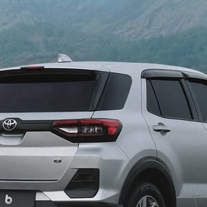 Silver Toyota Raize SUV parked on a dirt road in a lush, grassy field with mountains in the background under a cloudy sky.