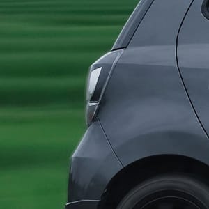 A dark gray hatchback drives past lush green rice paddies under a cloudy sky, conveying a sense of scenic travel.